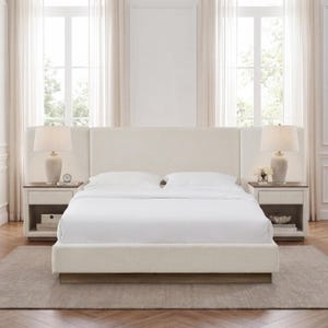 May include: A cream-colored upholstered bed frame with a tall, rectangular headboard and matching side wings. The bed is dressed in white linens and flanked by two wooden nightstands, each topped with a cream ceramic table lamp.
