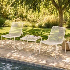 May include: White metal patio chairs and a small table with a mesh design. The chairs and table are on a stone patio next to a pool. A glass of water, sunglasses, and a book are on the table. A woven basket is on the right.