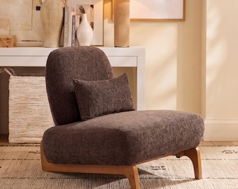 May include: A low-profile armless chair upholstered in dark brown textured fabric, featuring a rounded backrest and a matching rectangular lumbar pillow. The chair is supported by a light-toned wooden frame with four short, sturdy legs.