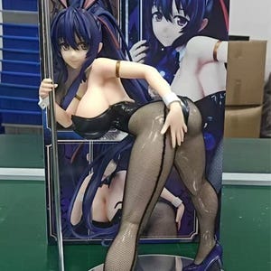 May include: A figurine of a woman in a black bodysuit, fishnet stockings, and high heels, with bunny ears. The figure is posed with one hand on a silver pole and is standing on a reflective base. The background features a box with a similar image.