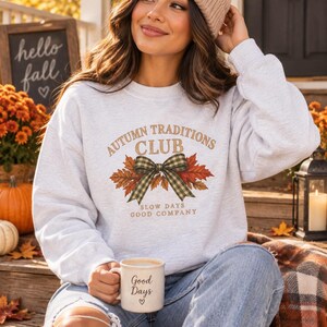 May include: A light gray crewneck sweatshirt featuring a graphic with the text "AUTUMN TRADITIONS CLUB" and "SLOW DAYS GOOD COMPANY" surrounding a green plaid bow and autumn leaves. The sweatshirt is paired with blue denim jeans and a beanie.