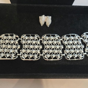 May include: A silver bracelet and matching earrings displayed on a black velvet surface. The bracelet has rectangular links with an openwork pattern. The earrings have a similar design.