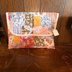 Puede incluir: Un bolso de mano de tela patchwork con un lazo blanco y una etiqueta en blanco. El bolso presenta una variedad de telas estampadas con flores, rayas y diseños en tonos rojos, azules, naranjas y blancos. La solapa es curva y está bordeada con tela blanca.
