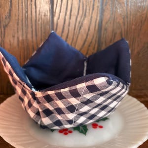 May include: A navy blue and white gingham bowl cozy sits on a white plate with a holly berry design. The bowl cozy is reversible, with a solid navy blue interior. The background is a wooden surface.