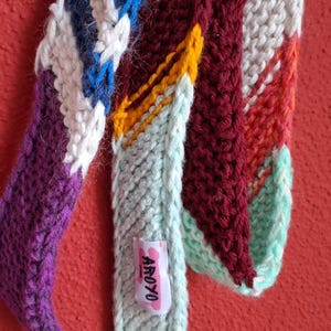Könnte beinhalten: Nahaufnahme eines bunten, gestrickten Schals mit verschiedenen Mustern und Farben. Der Schal hat Abschnitte in Weiß, Blau, Lila, Burgunderrot, Gelb, Orange und Mintgrün. Ein kleines Etikett mit dem Wort "AROYO" ist angebracht.