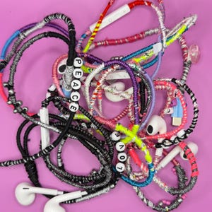 May include: A collection of colorful, handmade earbud holders with the words "PEACE" and "LOVE" spelled out in white letter beads. The holders are wrapped in various colors of thread, including black, pink, and blue, against a pink background.