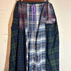 May include: A patchwork skirt made from various plaid flannel shirts. The skirt features a mix of blue, green, and red plaid patterns, with a drawstring waist and a button-down shirt front. The skirt is hanging on a black hanger.