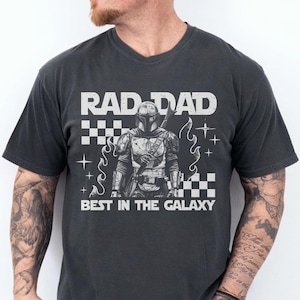 May include: Dark grey t-shirt featuring the text "RAD DAD BEST IN THE GALAXY" and a graphic of an armored character. The shirt has a crew neck and short sleeves. The design includes a checkered pattern and star accents.