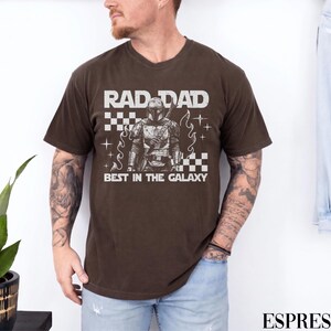 May include: Espresso-coloured t-shirt with the text "RAD DAD BEST IN THE GALAXY" in white. The graphic features a detailed illustration of a character in armour. The shirt is a v-neck style.