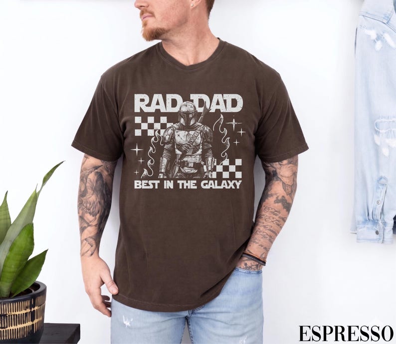 May include: Espresso-coloured t-shirt with the text "RAD DAD BEST IN THE GALAXY" in white. The graphic features a detailed illustration of a character in armour. The shirt is a v-neck style.