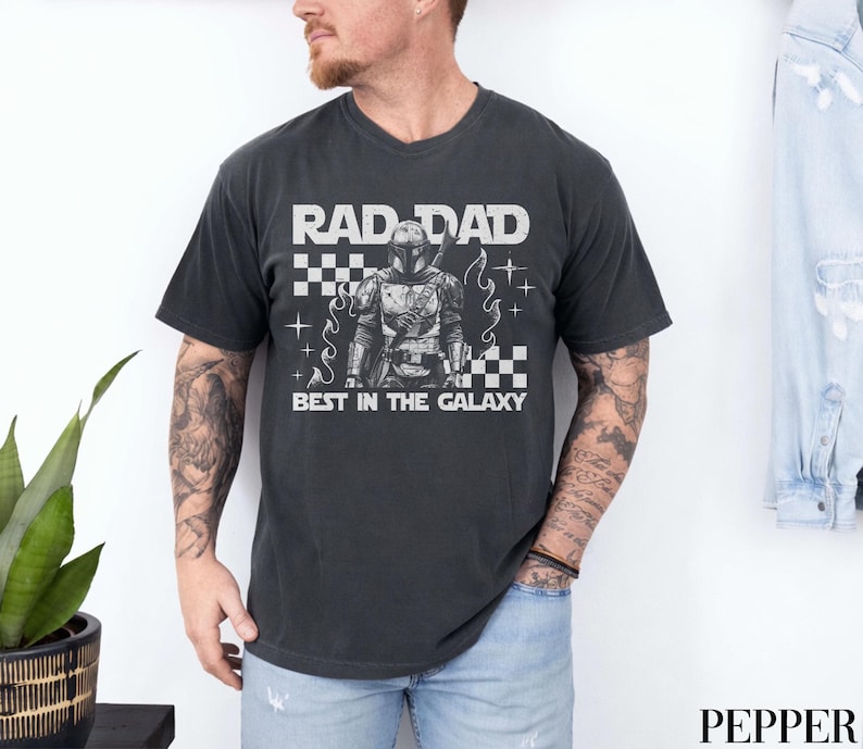 May include: Dark grey t-shirt featuring the text "RAD DAD BEST IN THE GALAXY" and a graphic of an armored character. The shirt has a crew neck and short sleeves. The design includes a checkered pattern and star accents.