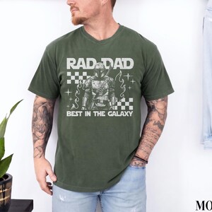 May include: An olive green t-shirt with the text "RAD DAD BEST IN THE GALAXY" in white. The graphic features a detailed illustration of a character in armor. The shirt has a crew neck and short sleeves.