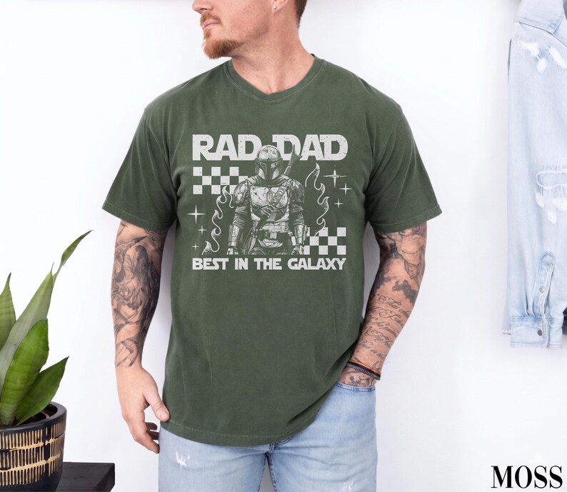 May include: An olive green t-shirt with the text "RAD DAD BEST IN THE GALAXY" in white. The graphic features a detailed illustration of a character in armor. The shirt has a crew neck and short sleeves.