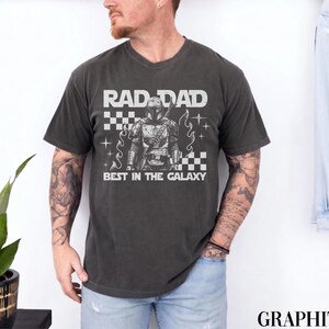 May include: Graphite grey t-shirt with the text "RAD DAD BEST IN THE GALAXY" in white. The graphic features a detailed illustration of a character in armour. The shirt has a classic crew neck and short sleeves.