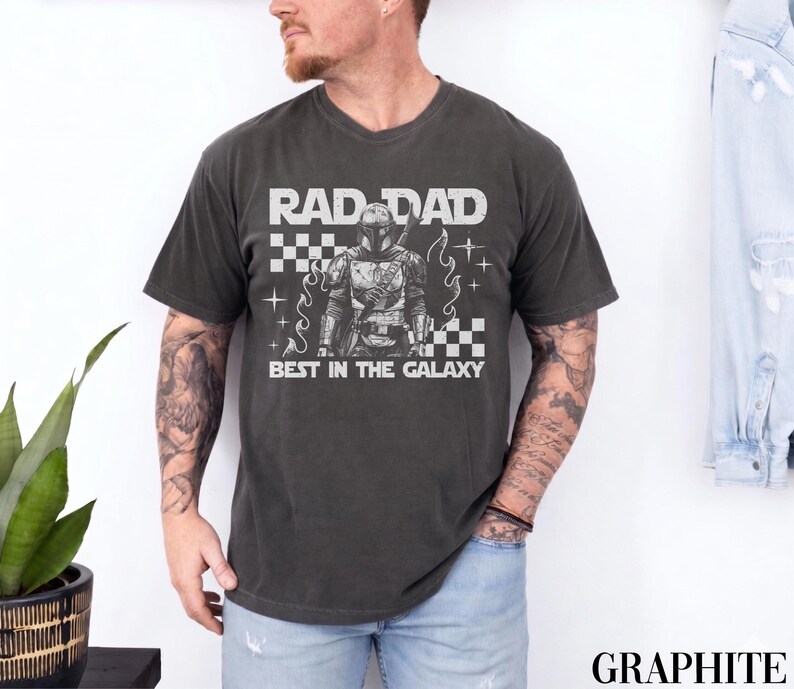 May include: Graphite grey t-shirt with the text "RAD DAD BEST IN THE GALAXY" in white. The graphic features a detailed illustration of a character in armour. The shirt has a classic crew neck and short sleeves.