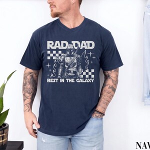 May include: Navy blue t-shirt with the text "RAD DAD BEST IN THE GALAXY" in white. The shirt features a graphic of a character in armor. The design includes a checkered pattern and star-like accents.