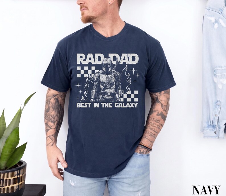 May include: Navy blue t-shirt with the text "RAD DAD BEST IN THE GALAXY" in white. The shirt features a graphic of a character in armor. The design includes a checkered pattern and star-like accents.