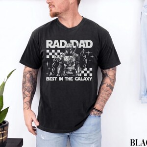 May include: Black t-shirt with the text "RAD DAD BEST IN THE GALAXY" in white. The graphic features a detailed illustration of a character in armour. The design includes checkered patterns and star-like accents.