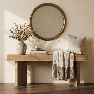 May include: A wooden bench with a round gold mirror hanging above it. A vase with dried plants, books, a small bowl, a candle, a patterned pillow, and a folded blanket are on the bench. The bench is made of light-colored wood.