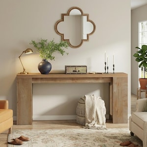 May include: A wooden console table with a decorative mirror, a blue vase with greenery, and a brass lamp. A small trunk and candlesticks are on the table. A woven pouf and a blanket are below the table. The room has a neutral color palette.