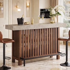 May include: A modern home bar with a concrete countertop and a slatted wooden front. The bar features a black ice bucket, a gold cocktail shaker, and two brown leather bar stools. A vase of white flowers and two champagne flutes are on the bar.