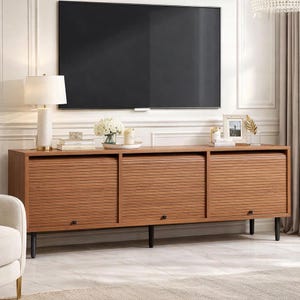May include: A wooden media console with three doors featuring a slatted design. The console is brown with black legs and handles. A large black television is mounted above the console. A lamp, flowers, and decorative items are on top of the console.