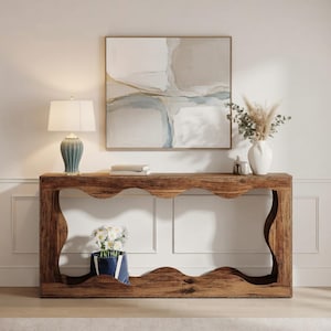 May include: A rustic wooden console table with a unique wavy edge design. The table is adorned with a blue ceramic lamp, books, a white vase with dried flowers, and a blue tote bag with white flowers. A framed abstract painting hangs above.