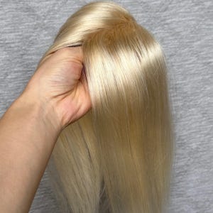 May include: A hair topper featuring straight, light platinum blonde hair with a visible monofilament base at the crown. The hair is smooth and sleek, showcasing a natural-looking part where the strands meet the scalp-colored base material.