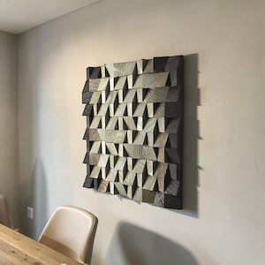 May include: Geometric wooden wall art with a textured, layered design. The artwork features a repeating pattern of angled blocks in shades of gray, brown, and natural wood tones. The piece is mounted on a neutral-colored wall, creating a modern, abstract aesthetic.