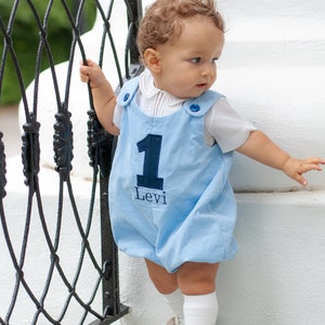 May include: A baby wearing a light blue romper with the number 1 and the name "Levi" in navy blue. The romper has blue button details and is paired with a white collared shirt, white socks, and white shoes. The baby is standing near a black metal fence.