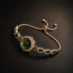 May include: A gold-tone bracelet watch featuring a round green dial with gold numerals, surrounded by a halo of clear crystals. The band consists of alternating oval green stones and clear crystal accents, finished with an adjustable sliding clasp.