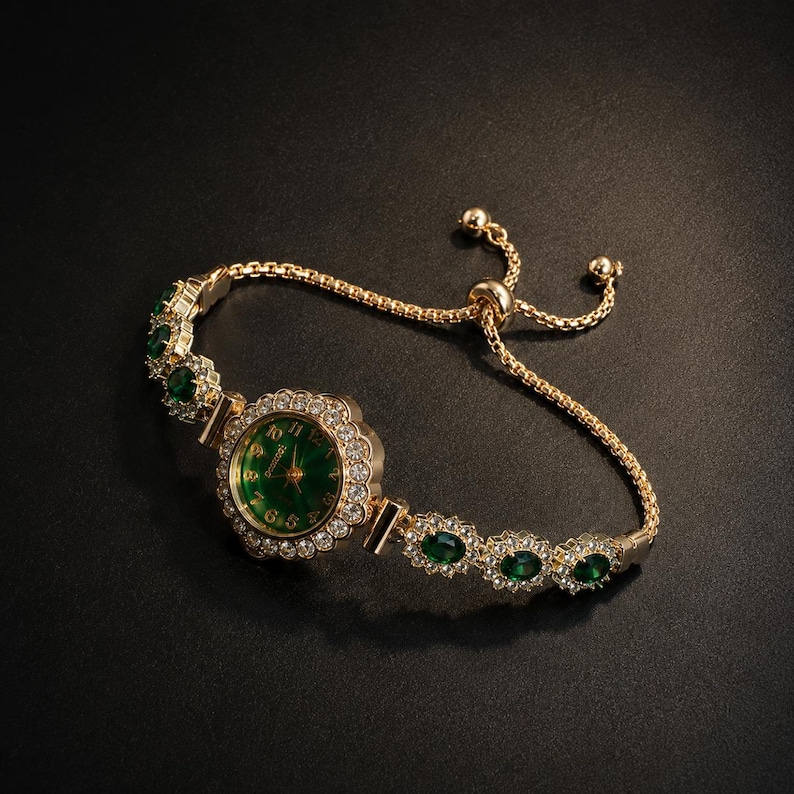 May include: A gold-tone bracelet watch featuring a round green dial with gold numerals, surrounded by a halo of clear crystals. The band consists of alternating oval green stones and clear crystal accents, finished with an adjustable sliding clasp.