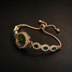 May include: A gold-tone bracelet watch featuring a round green dial with gold numerals, surrounded by a halo of clear crystals. The band consists of a box chain with three oval green stones set in crystal-encrusted frames and an adjustable slider.