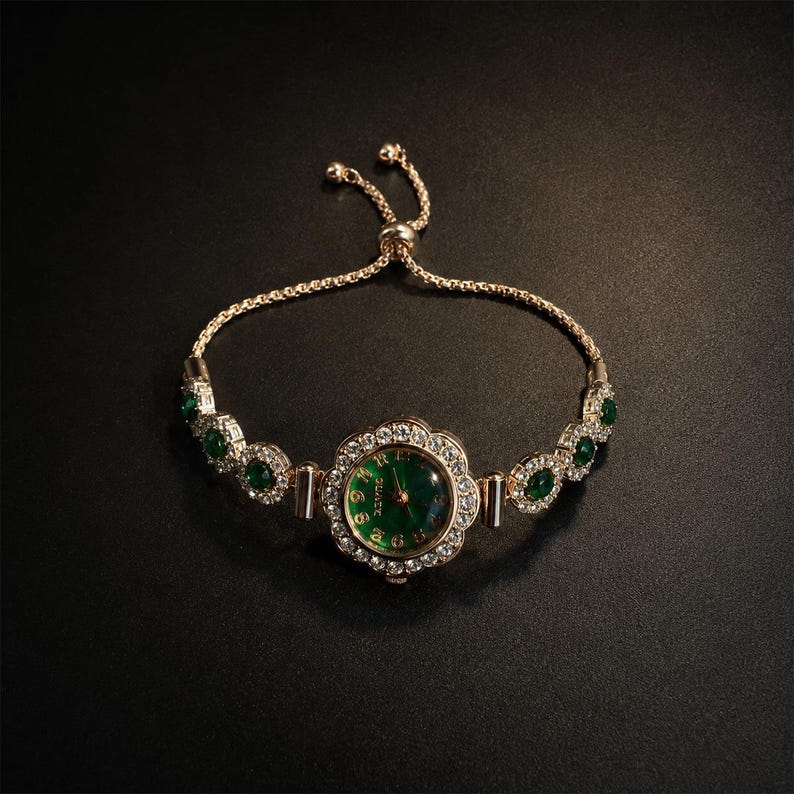 May include: A gold-tone bracelet watch featuring a round, emerald green dial surrounded by a floral-shaped bezel set with clear crystals. The band consists of alternating clear crystal and emerald green stone links with an adjustable sliding clasp.