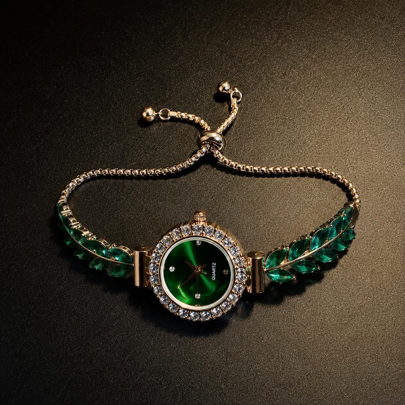May include: A gold-tone quartz watch featuring a round green dial surrounded by a bezel of clear crystals. The watch band is composed of gold-tone metal links adorned with marquise-cut green stones and includes an adjustable sliding clasp.