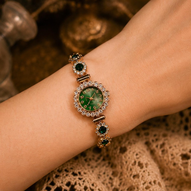 May include: A gold-tone analog watch featuring a round green dial with gold numerals. The bezel and bracelet are embellished with clear white crystals and circular green stones, creating a floral-inspired link design around the wrist.