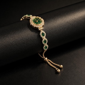 May include: A gold-tone quartz watch with a round green face, surrounded by a halo of clear crystals. The bracelet features alternating oval green stones and clear crystal accents, finished with a sliding adjustable chain and two gold-tone beads.
