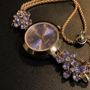 May include: A rose gold-tone quartz watch featuring a round purple dial with gold-tone numerals. The watch band is a rose gold-tone chain adorned with two clusters of light purple, petal-shaped crystals on each side of the watch face.