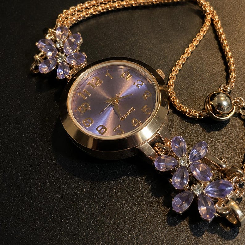 May include: A rose gold-tone quartz watch featuring a round purple dial with gold-tone numerals. The watch band is a rose gold-tone chain adorned with two clusters of light purple, petal-shaped crystals on each side of the watch face.