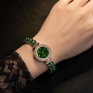 May include: A gold-tone quartz watch featuring a round green dial surrounded by a ring of clear crystals. The watch band is composed of a series of marquise-cut green stones arranged in a leaf-like pattern, creating a decorative bracelet design.