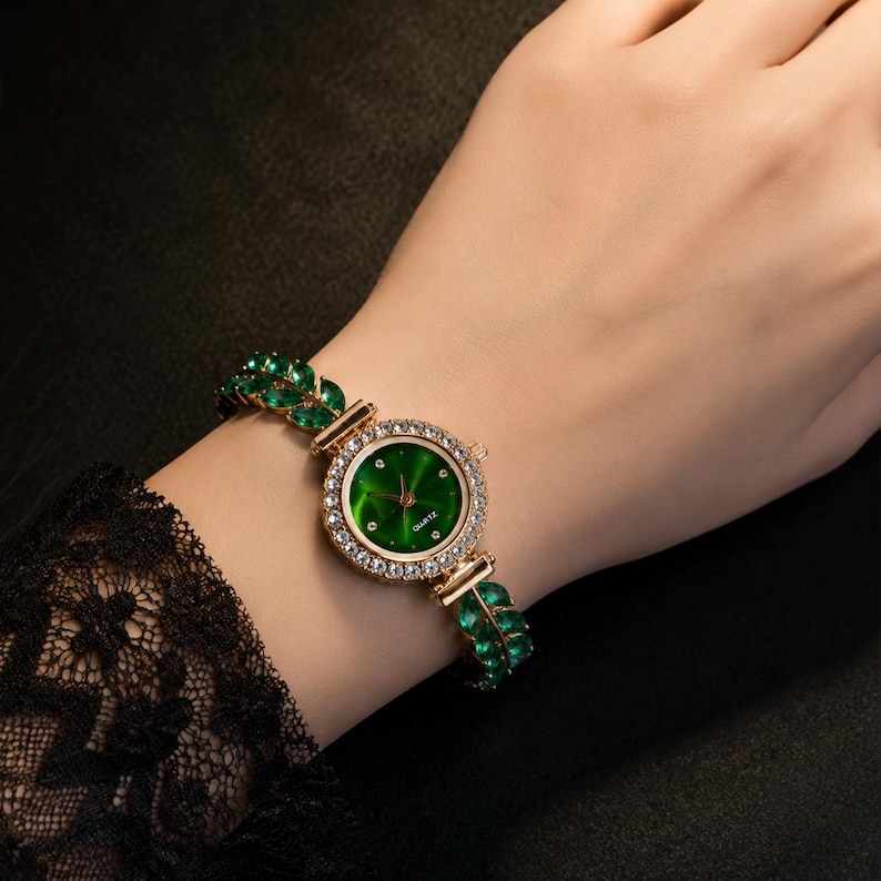 May include: A gold-tone quartz watch featuring a round green dial surrounded by a ring of clear crystals. The watch band is composed of a series of marquise-cut green stones arranged in a leaf-like pattern, creating a decorative bracelet design.
