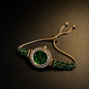 May include: A gold-tone quartz watch with a round green dial featuring small crystal hour markers. The bezel is encrusted with clear stones. The bracelet is a gold-tone chain with green marquise-cut stones and an adjustable sliding clasp.