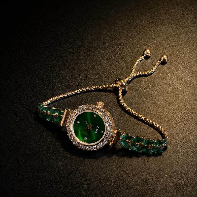 May include: A gold-tone quartz watch with a round green dial featuring small crystal hour markers. The bezel is encrusted with clear stones. The bracelet is a gold-tone chain with green marquise-cut stones and an adjustable sliding clasp.