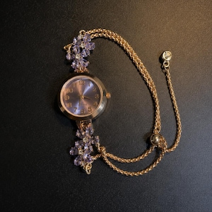 May include: A gold-tone quartz watch featuring a round face with a purple dial and gold numerals. The watch band is a double gold chain adorned with two floral accents made of light purple petals and clear stones, plus an adjustable slider.