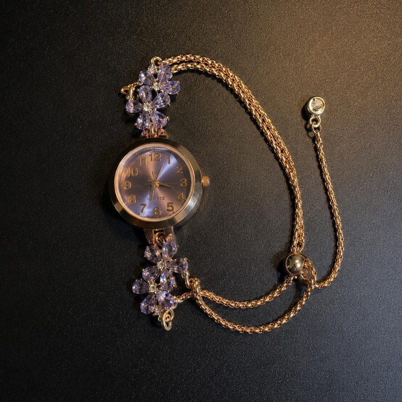 May include: A gold-tone quartz watch featuring a round face with a purple dial and gold numerals. The watch band is a double gold chain adorned with two floral accents made of light purple petals and clear stones, plus an adjustable slider.
