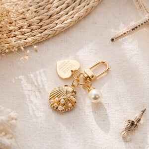 May include: A gold-toned metal keychain featuring a heart-shaped charm with the engraved text 'JE T'AIME', a textured seashell charm adorned with small white pearls, and a single round white pearl bead attached to a lobster claw clasp.