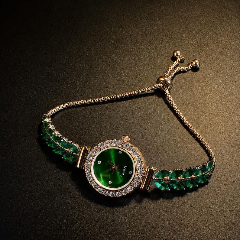 May include: A gold-tone quartz watch featuring a round green dial with small crystal markers. The bezel is encrusted with clear crystals, and the bracelet consists of gold-tone chain links accented with marquise-cut green stones on each side.