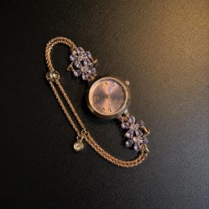 May include: A rose gold-tone quartz watch featuring a round face with gold numerals and a metallic dial. The bracelet consists of two delicate chains adorned with two floral-shaped clusters of light purple crystals and a single round crystal charm.