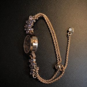 May include: A gold-tone wristwatch featuring a round dark face with numerals, connected to a double-strand chain bracelet. The watch band is accented with clusters of light purple floral-shaped stones and includes a sliding adjustable bead clasp.