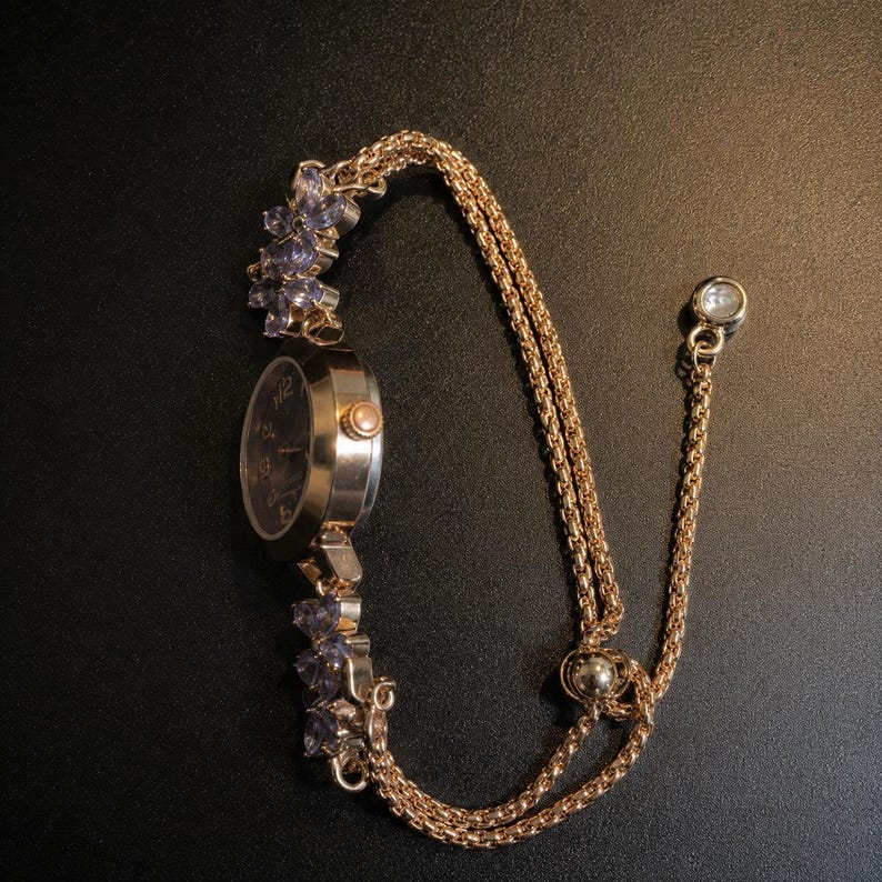 May include: A gold-tone wristwatch featuring a round dark face with numerals, connected to a double-strand chain bracelet. The watch band is accented with clusters of light purple floral-shaped stones and includes a sliding adjustable bead clasp.